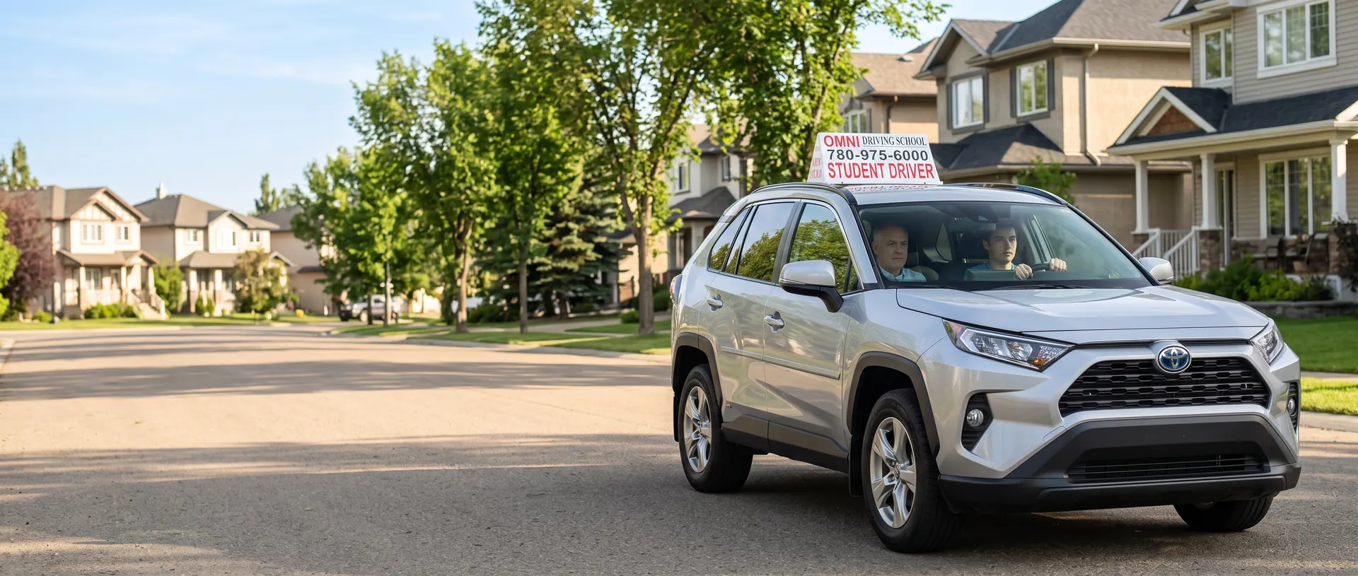 A student drives the Omni Driving School car on a quiet street of houses, the instructor in the passenger seat